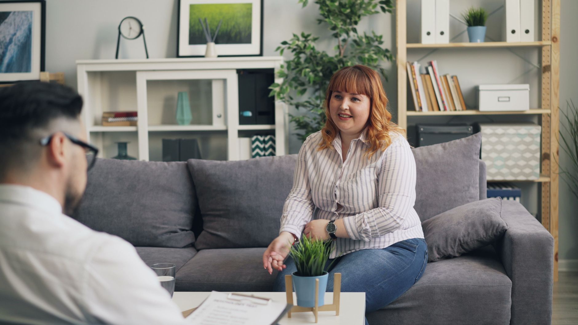 A woman sitting on a gray couch, engaging in conversation with a man who is partially visible. She has curly red hair and is wearing a striped blouse. A small potted plant and a glass of water are on the table in front of her.