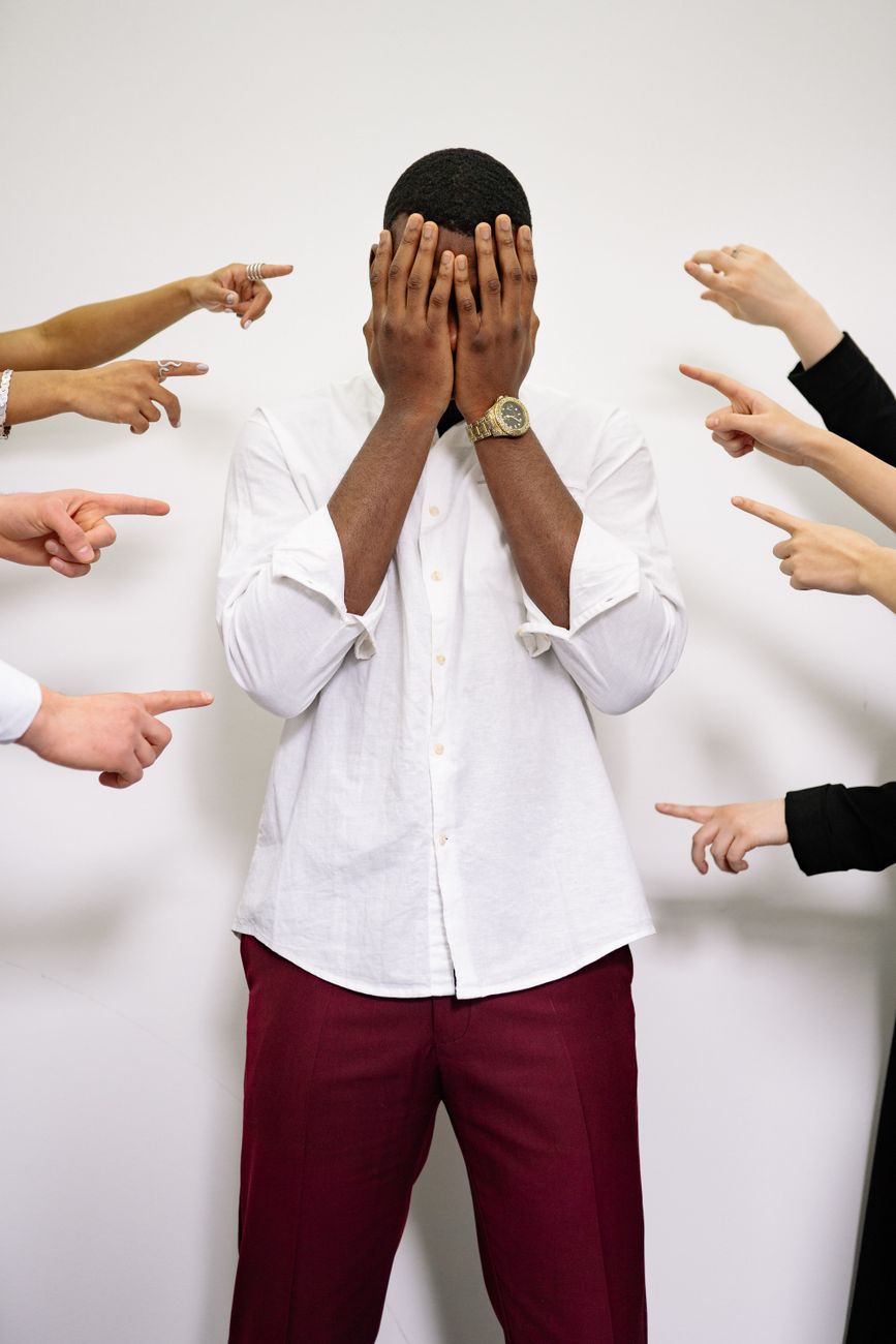 A man in a white shirt and maroon pants stands facing the camera with his hands covering his face, while several hands point at him from different angles.