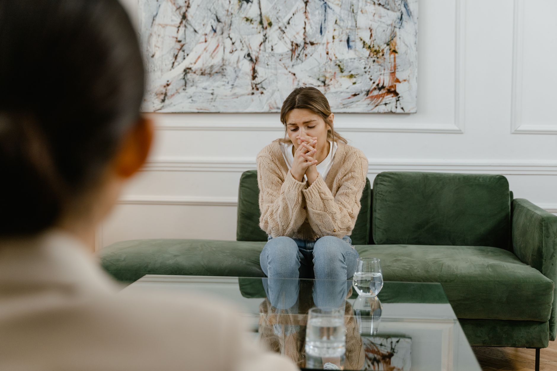 A woman sitting on a green couch, looking distressed with her hands clasped together, while another person, partially out of focus, sits across from her. A glass of water is on the table between them.